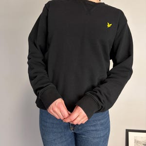 Puede incluir: Una sudadera negra de manga larga con cuello redondo y un detalle acanalado en forma de V en el cuello. La prenda tiene pu&ntilde;os y dobladillo acanalados, con un peque&ntilde;o logotipo de &aacute;guila bordado en amarillo en la parte superior izquierda.