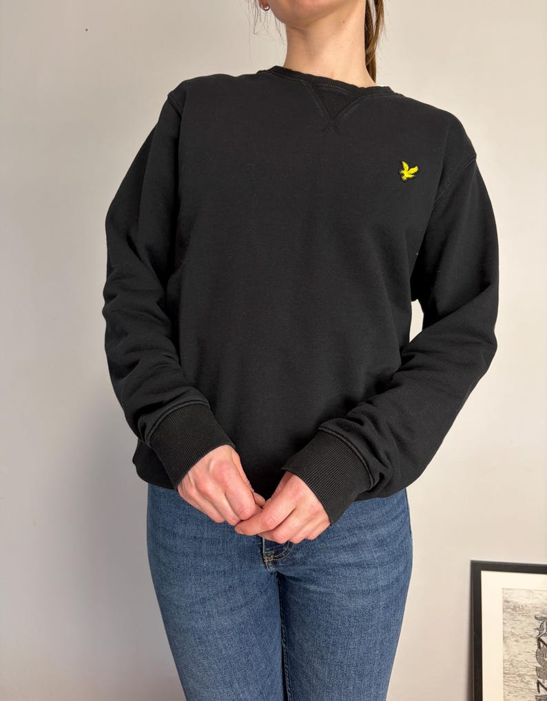 Puede incluir: Una sudadera negra de manga larga con cuello redondo y un detalle acanalado en forma de V en el cuello. La prenda tiene pu&ntilde;os y dobladillo acanalados, con un peque&ntilde;o logotipo de &aacute;guila bordado en amarillo en la parte superior izquierda.
