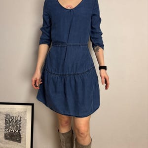 Puede incluir: Vestido vaquero azul oscuro con cuello en V y mangas largas. El vestido tiene cintura entallada y falda escalonada. La modelo lleva botas grises hasta la rodilla. Se ve un estampado enmarcado con el texto "Don't Make Sense Make".