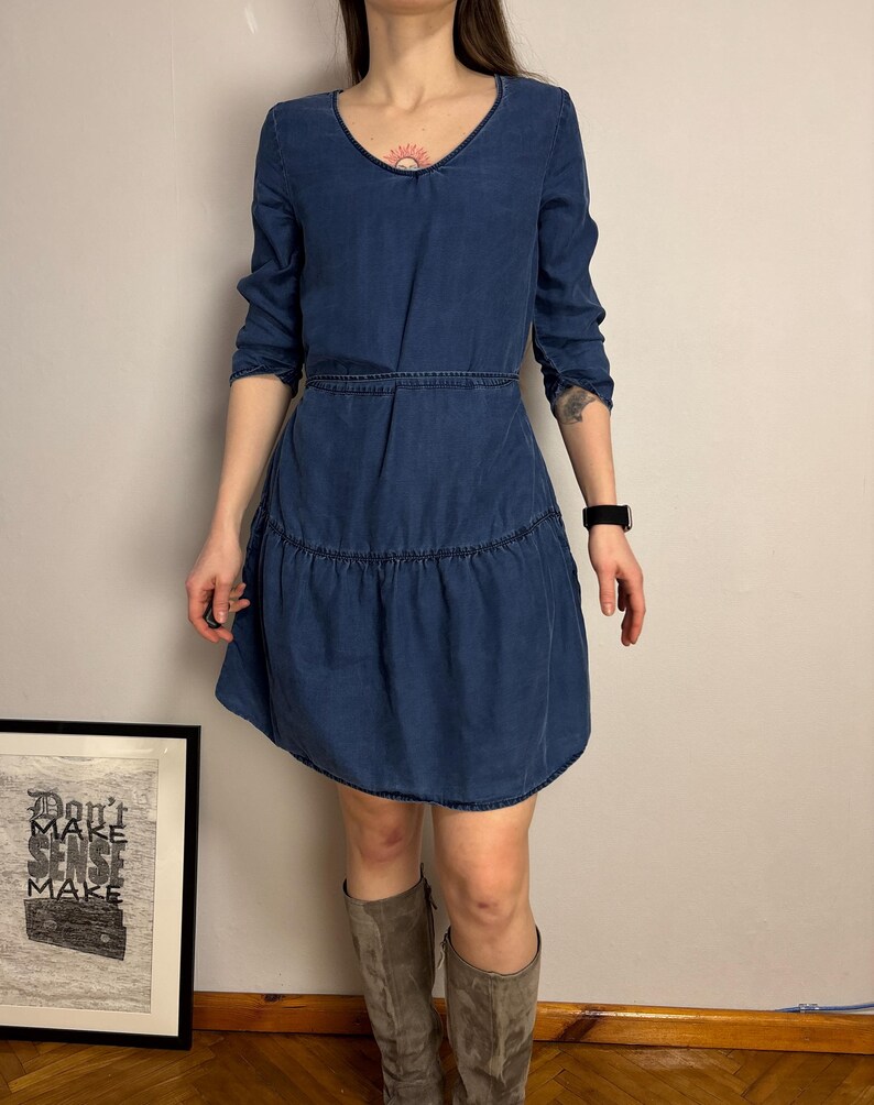 Puede incluir: Vestido vaquero azul oscuro con cuello en V y mangas largas. El vestido tiene cintura entallada y falda escalonada. La modelo lleva botas grises hasta la rodilla. Se ve un estampado enmarcado con el texto "Don't Make Sense Make".