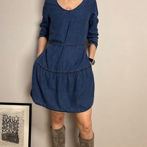 Puede incluir: Un vestido de mezclilla azul con cuello en V y mangas largas. El vestido presenta una falda escalonada y bolsillos. La modelo lleva botas grises. Se ve una impresi&oacute;n enmarcada con el texto "Don't Make Sense Make".