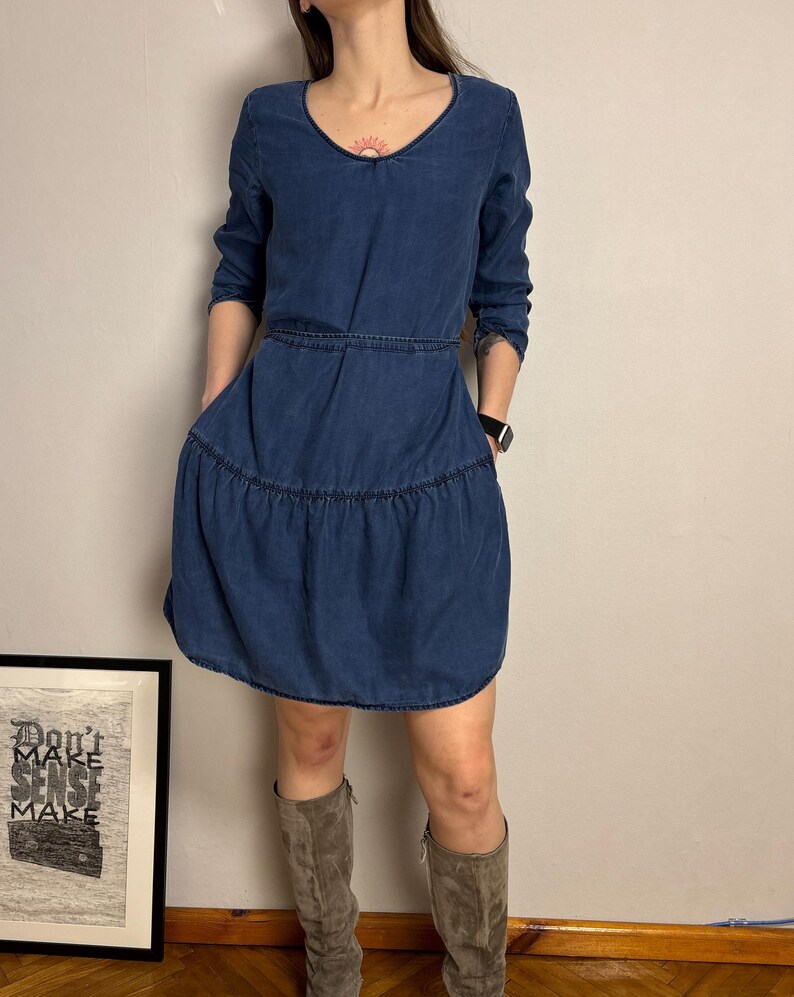 Puede incluir: Un vestido de mezclilla azul con cuello en V y mangas largas. El vestido presenta una falda escalonada y bolsillos. La modelo lleva botas grises. Se ve una impresi&oacute;n enmarcada con el texto "Don't Make Sense Make".