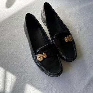 Może przedstawiać: Para czarnych sk&oacute;rzanych mokasyn&oacute;w ze złotymi elementami. Buty mają klasyczny design z zaokrąglonym noskiem i niskim obcasem. Mokasyny są prezentowane na białej powierzchni.