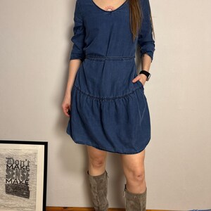 Puede incluir: Vestido de mezclilla azul con cuello en V y mangas largas. El vestido presenta una cintura entallada y una falda escalonada. La modelo lleva botas de ante grises. Una obra de arte enmarcada con el texto "Don't Make Sense Make" es visible en el fondo.