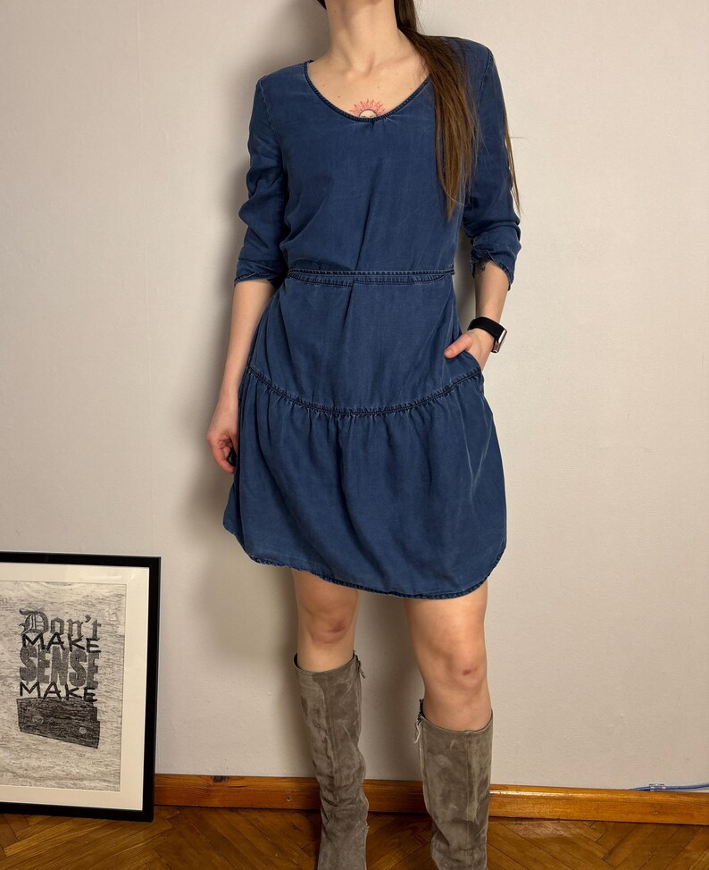 Puede incluir: Vestido de mezclilla azul con cuello en V y mangas largas. El vestido presenta una cintura entallada y una falda escalonada. La modelo lleva botas de ante grises. Una obra de arte enmarcada con el texto "Don't Make Sense Make" es visible en el fondo.