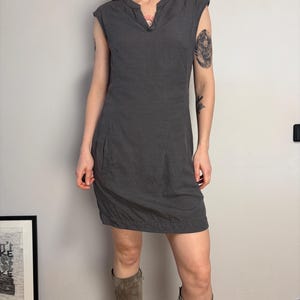 May include: A dark gray sleeveless dress featuring a notched neckline, a fitted bodice, and a straight skirt that hits above the knee. The dress includes visible side pockets and a subtle horizontal seam detail along the bottom hemline.