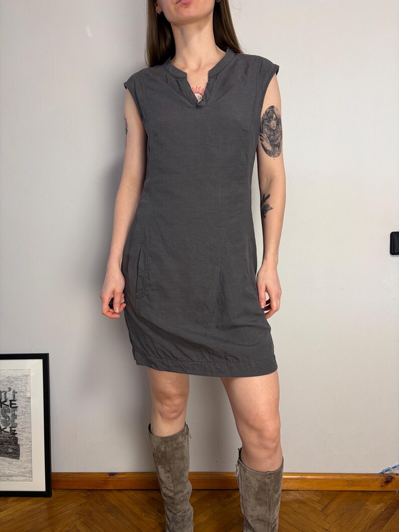 May include: A dark gray sleeveless dress featuring a notched neckline, a fitted bodice, and a straight skirt that hits above the knee. The dress includes visible side pockets and a subtle horizontal seam detail along the bottom hemline.