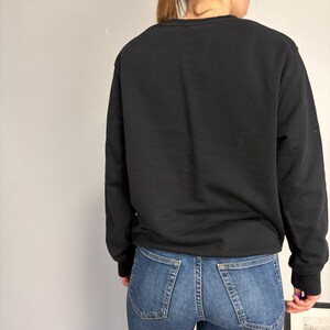 Puede incluir: Una sudadera negra de manga larga con cuello redondo combinada con pantalones vaqueros azules. La sudadera presenta un dise&ntilde;o liso y sencillo con pu&ntilde;os y cuello acanalados, mientras que los vaqueros tienen un estilo cl&aacute;sico de cinco bolsillos con costuras visibles.