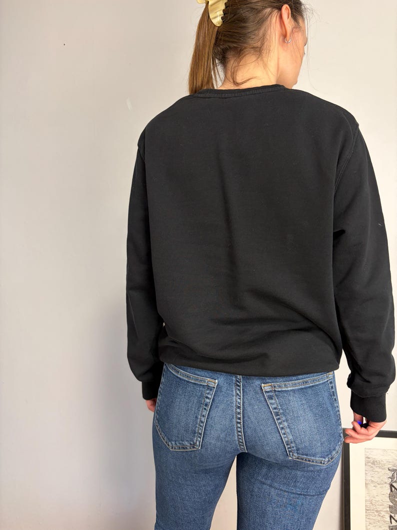 Puede incluir: Una sudadera negra de manga larga con cuello redondo combinada con pantalones vaqueros azules. La sudadera presenta un dise&ntilde;o liso y sencillo con pu&ntilde;os y cuello acanalados, mientras que los vaqueros tienen un estilo cl&aacute;sico de cinco bolsillos con costuras visibles.