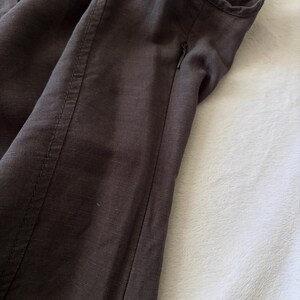 May include: A dark brown garment featuring a visible side seam with reinforced stitching and a small, integrated zipper closure. The fabric displays a subtle, textured weave pattern throughout the surface, highlighting the construction details.