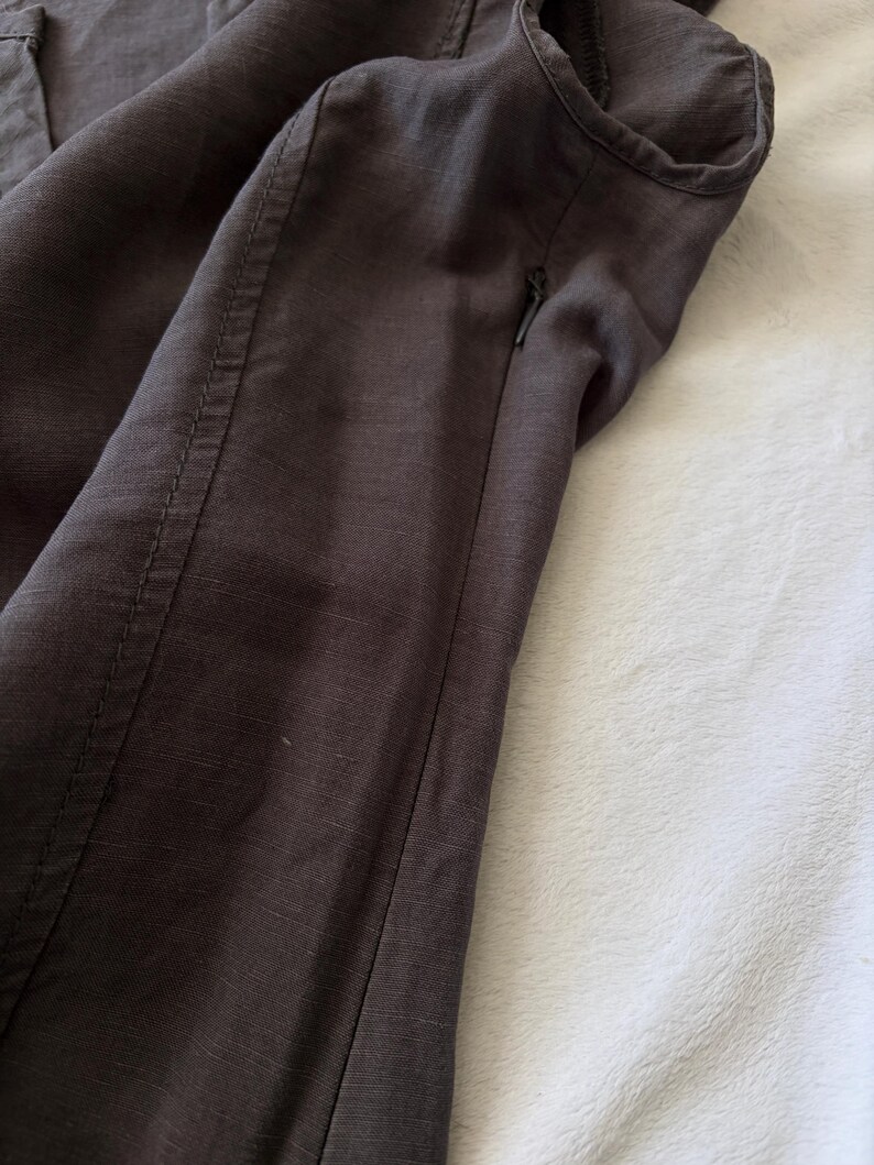 May include: A dark brown garment featuring a visible side seam with reinforced stitching and a small, integrated zipper closure. The fabric displays a subtle, textured weave pattern throughout the surface, highlighting the construction details.