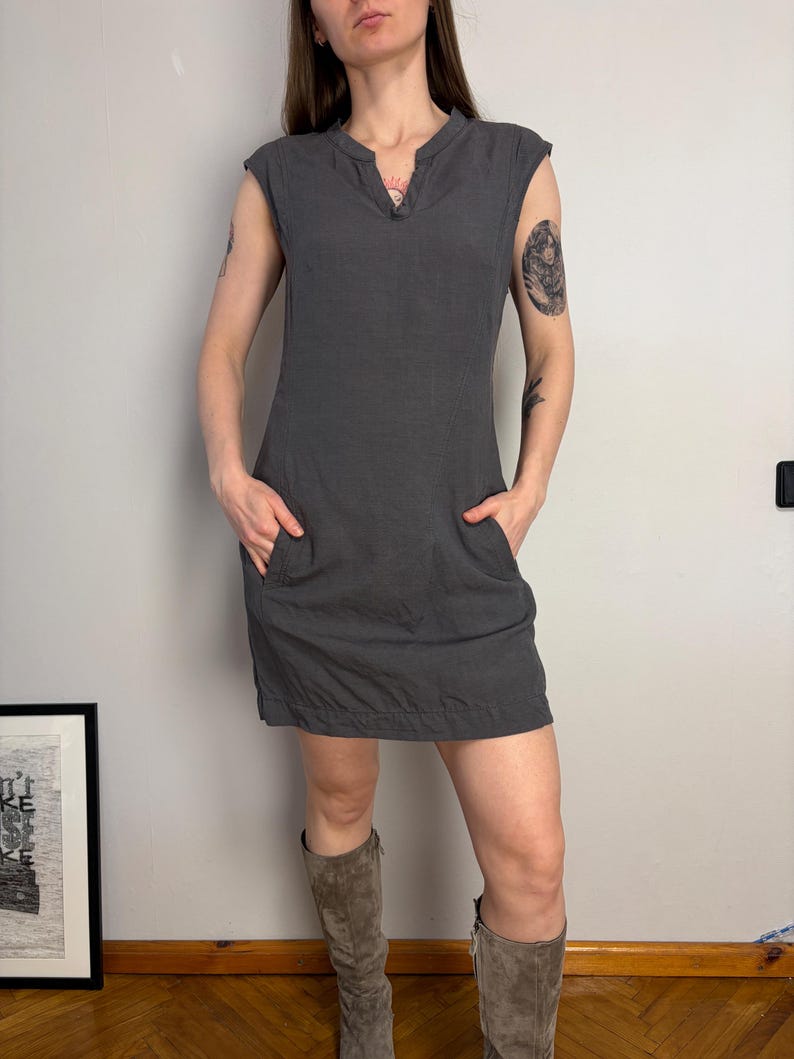 May include: A dark gray sleeveless mini dress featuring a notched V-neckline, a fitted bodice, and two side pockets. The dress has a straight hemline and a simple, clean construction, providing a versatile look that falls above the knee.