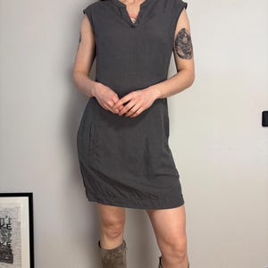 May include: A charcoal gray sleeveless dress featuring a notched neckline, cap sleeves, and a straight hemline. The dress has a simple, unadorned design with a visible seam running down the front and side pockets integrated into the skirt portion.