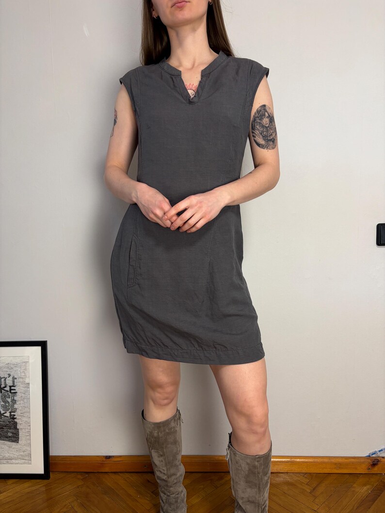 May include: A charcoal gray sleeveless dress featuring a notched neckline, cap sleeves, and a straight hemline. The dress has a simple, unadorned design with a visible seam running down the front and side pockets integrated into the skirt portion.