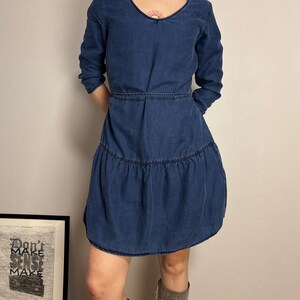 Puede incluir: Un vestido de mezclilla azul con cuello en V y mangas largas. El vestido tiene cintura entallada y falda escalonada. La modelo lleva botas grises. En el fondo se ve una impresi&oacute;n enmarcada con el texto "Don't Make Sense Make".