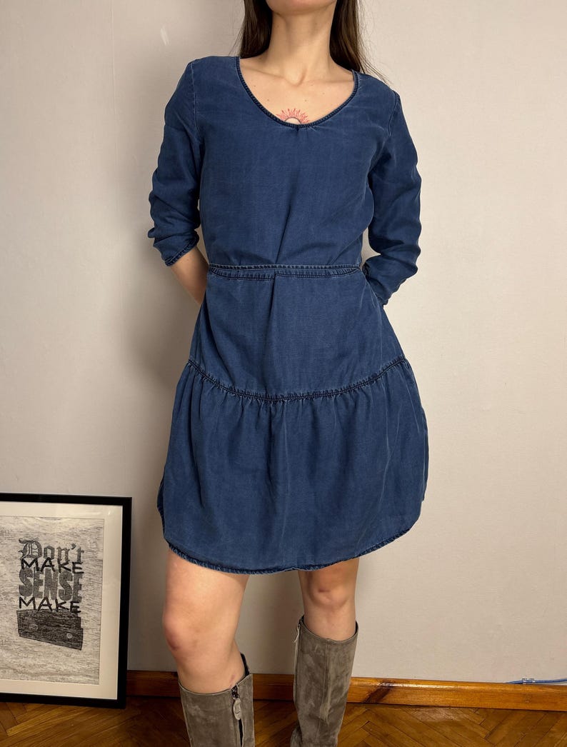 Puede incluir: Un vestido de mezclilla azul con cuello en V y mangas largas. El vestido tiene cintura entallada y falda escalonada. La modelo lleva botas grises. En el fondo se ve una impresi&oacute;n enmarcada con el texto "Don't Make Sense Make".