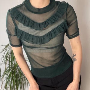 May include: A sheer, dark green top with short sleeves and a high neckline. The top features three rows of ruffled trim across the chest. The model is wearing black trousers. The top is made of a see-through material.