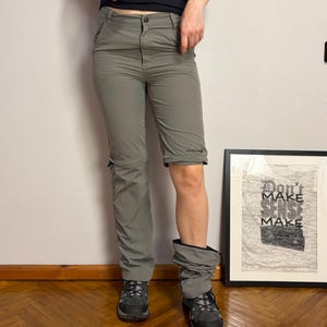 Puede incluir: Pantalones de senderismo convertibles de color verde oliva con cierre de botón, trabillas para cinturón y secciones con cremallera por encima de la rodilla que permiten separar la parte inferior de las piernas. Se combinan con botas de montaña gris oscuro.