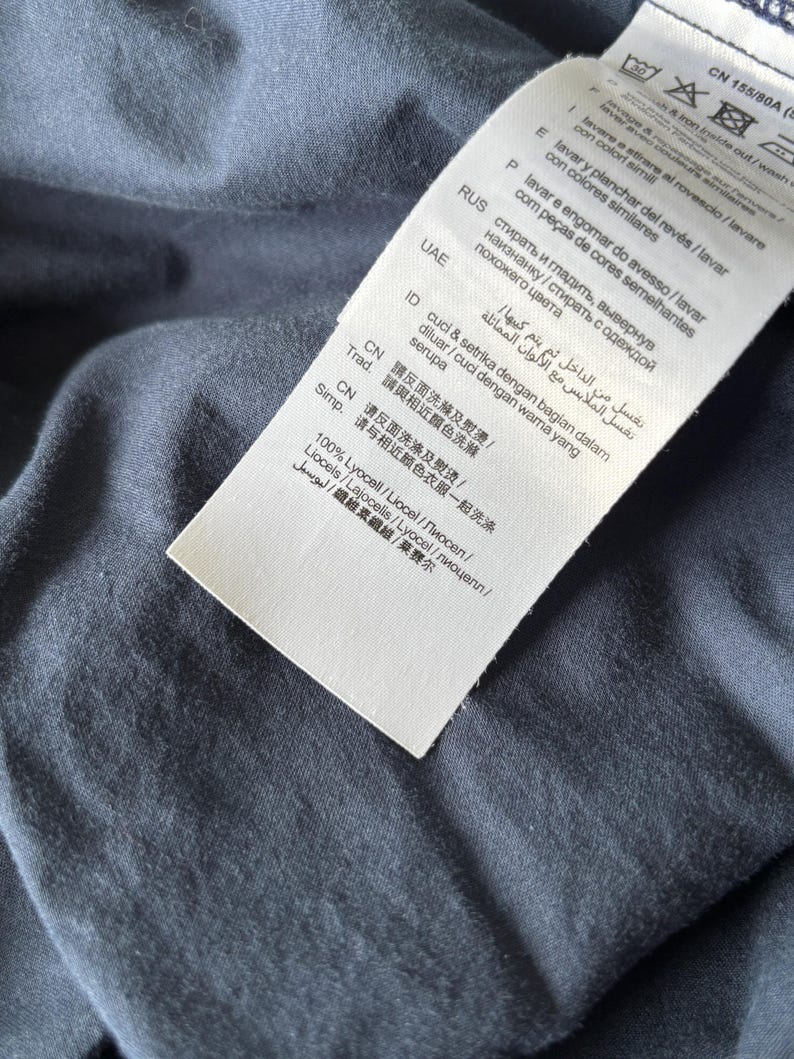 Puede incluir: Primer plano de una tela azul marino con una etiqueta blanca adjunta. La etiqueta contiene instrucciones de lavado en varios idiomas, incluido el espa&ntilde;ol. La tela parece suave y ligeramente texturizada.