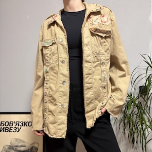 May include: Tan jacket with floral and star embellishments. The jacket has a button-up front, multiple pockets, and decorative patches. The edges of the jacket have a frayed design. The jacket is worn over a black top and black pants.