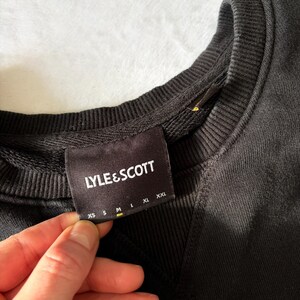 Puede incluir: Una sudadera negra con cuello redondo acanalado y un detalle de costura triangular debajo del cuello. Una etiqueta de tela negra est&aacute; cosida en el interior del cuello, mostrando el texto blanco LYLE & SCOTT sobre una tabla de tallas XS, S, M, L, XL y XXL.