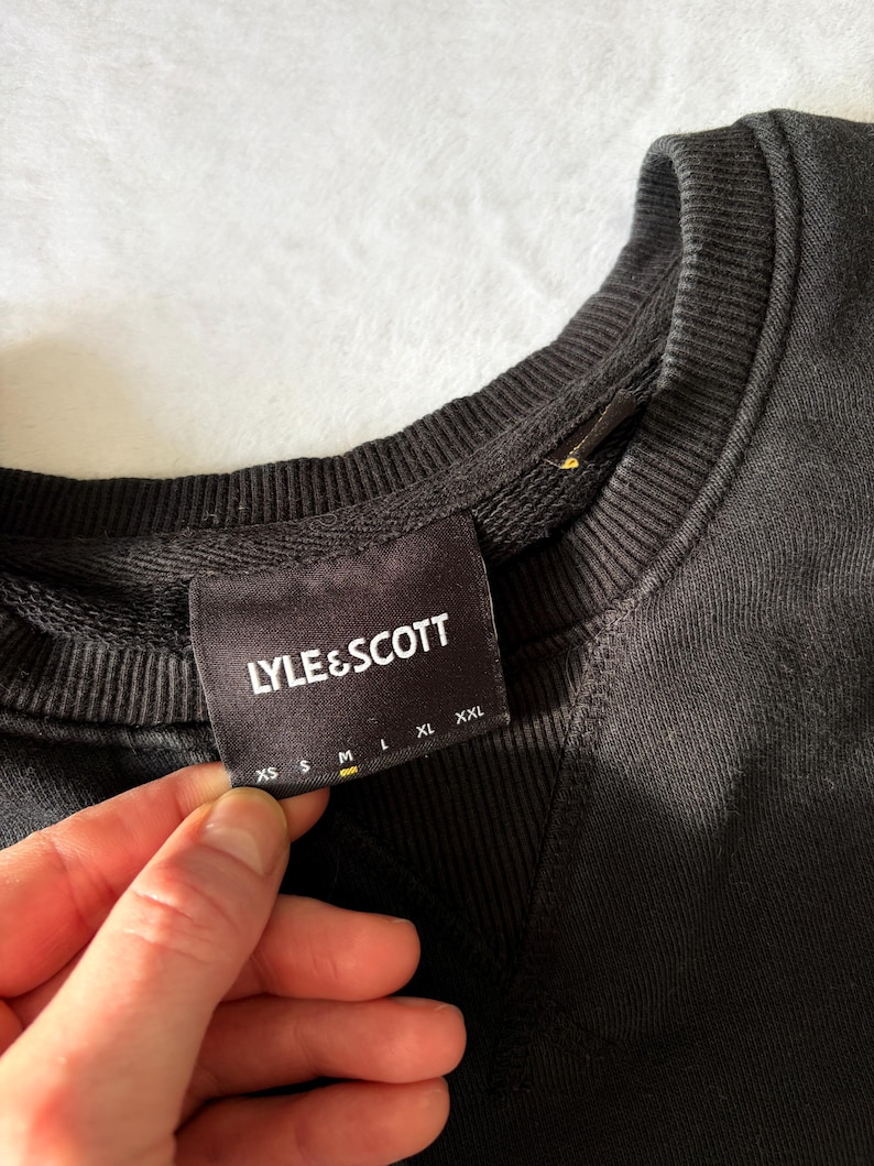 Puede incluir: Una sudadera negra con cuello redondo acanalado y un detalle de costura triangular debajo del cuello. Una etiqueta de tela negra est&aacute; cosida en el interior del cuello, mostrando el texto blanco LYLE & SCOTT sobre una tabla de tallas XS, S, M, L, XL y XXL.