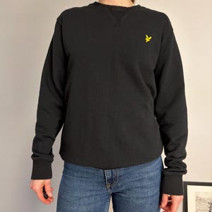 Puede incluir: Una sudadera negra de manga larga con cuello redondo y un detalle de inserci&oacute;n en V acanalado. La prenda presenta un peque&ntilde;o logotipo de &aacute;guila bordado en amarillo en el pecho izquierdo y pu&ntilde;os y dobladillo acanalados, combinada con pantalones vaqueros azules.