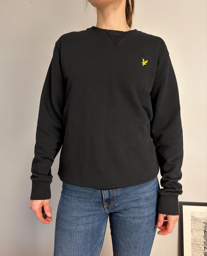 Puede incluir: Una sudadera negra de manga larga con cuello redondo y un detalle de inserci&oacute;n en V acanalado. La prenda presenta un peque&ntilde;o logotipo de &aacute;guila bordado en amarillo en el pecho izquierdo y pu&ntilde;os y dobladillo acanalados, combinada con pantalones vaqueros azules.
