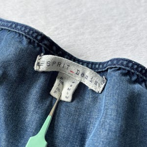 Puede incluir: Primer plano de una prenda vaquera azul con una etiqueta que dice "ESPRIT Denim". La etiqueta tambi&eacute;n muestra informaci&oacute;n sobre la talla. Una herramienta verde claro sostiene la etiqueta.