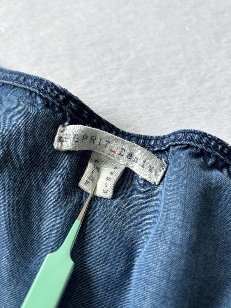 Puede incluir: Primer plano de una prenda vaquera azul con una etiqueta que dice "ESPRIT Denim". La etiqueta tambi&eacute;n muestra informaci&oacute;n sobre la talla. Una herramienta verde claro sostiene la etiqueta.