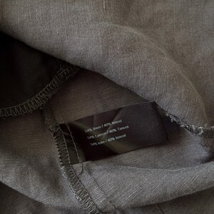 May include: A dark gray fabric garment featuring a black interior care label. The label displays the text 54% linen / 46% tencel, 54% Leinen / 46% Tencel, and 54% toile / 46% tencel. The material has a visible woven texture and stitched seams.