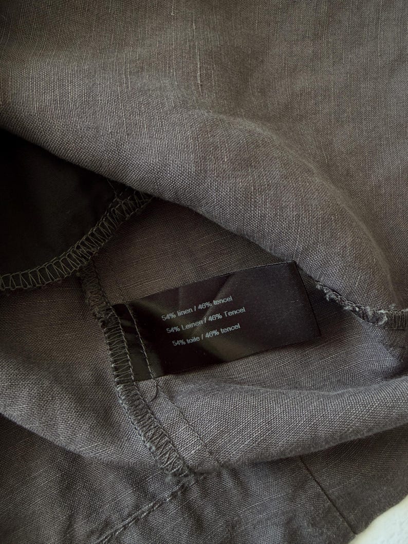 May include: A dark gray fabric garment featuring a black interior care label. The label displays the text 54% linen / 46% tencel, 54% Leinen / 46% Tencel, and 54% toile / 46% tencel. The material has a visible woven texture and stitched seams.