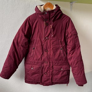 May include: A deep burgundy hooded parka featuring a thick cream-colored sherpa lining, a front button closure with a high collar, two large lower flap pockets with snap buttons, two vertical chest pockets, and a small utility pocket on the sleeve.