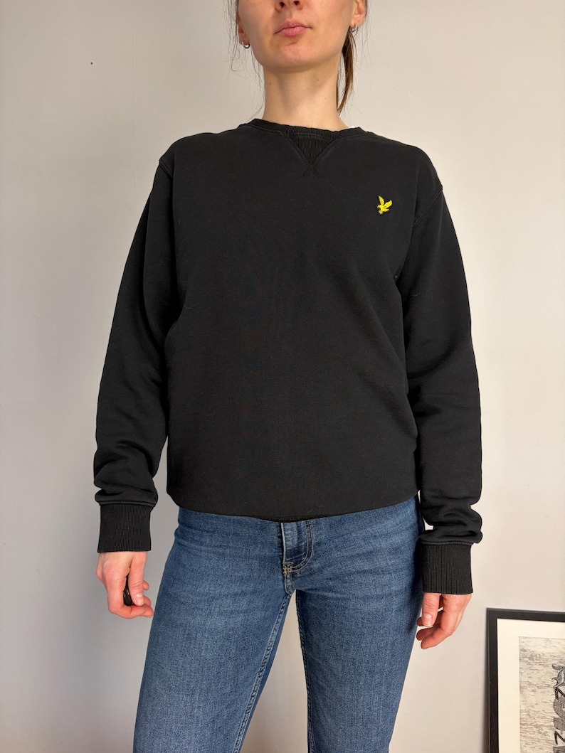 Puede incluir: Una sudadera negra de manga larga con cuello redondo, que presenta un cuello acanalado con un detalle de costura en forma de V en el escote y un peque&ntilde;o logotipo de p&aacute;jaro bordado en amarillo en el pecho izquierdo, junto con vaqueros azules.