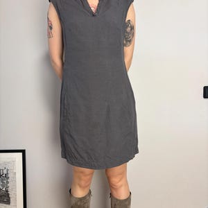 May include: A charcoal gray sleeveless dress featuring a split V-neckline, cap sleeves, and a straight silhouette that falls above the knee. The dress includes visible vertical seam detailing along the front and side pockets integrated into the design.