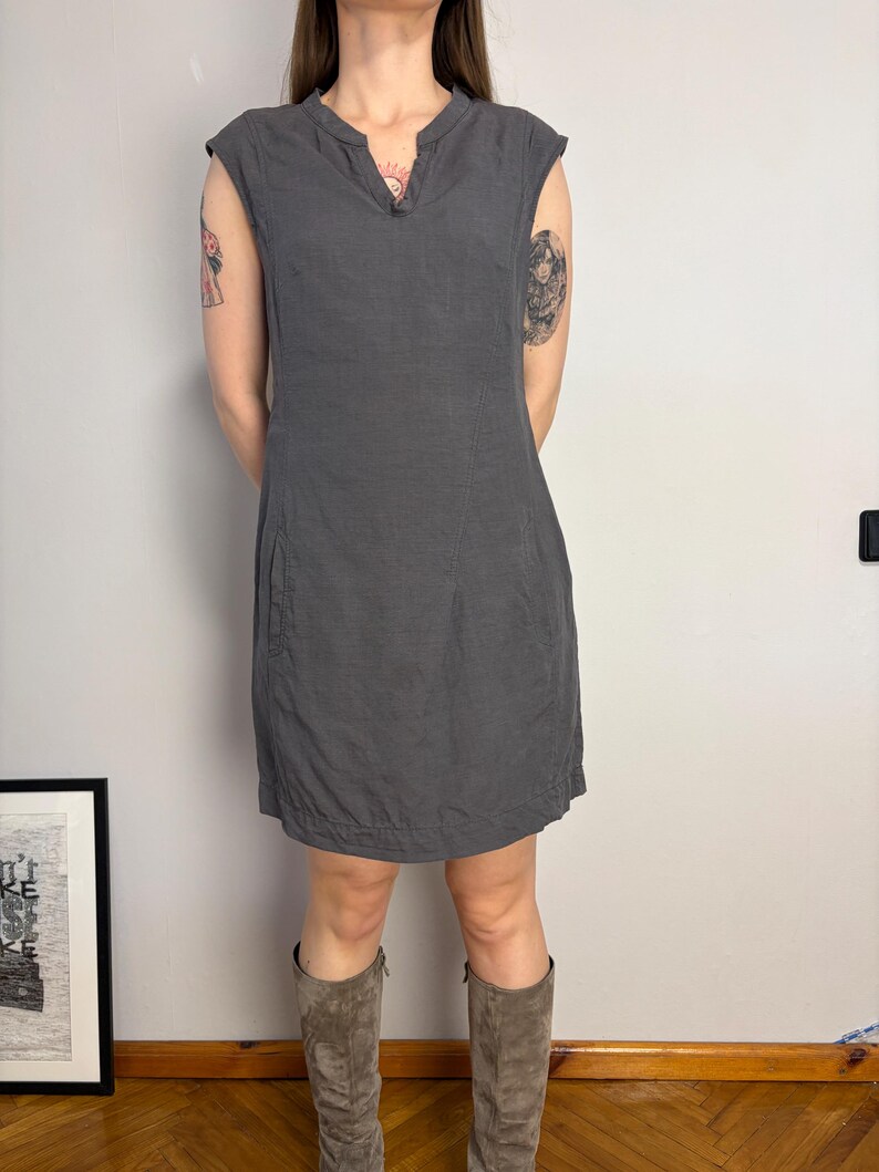May include: A charcoal gray sleeveless dress featuring a split V-neckline, cap sleeves, and a straight silhouette that falls above the knee. The dress includes visible vertical seam detailing along the front and side pockets integrated into the design.