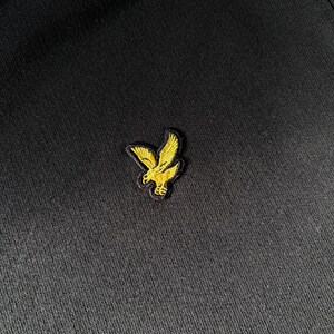 Puede incluir: Una prenda de tela negra con un peque&ntilde;o logotipo bordado de un &aacute;guila de color amarillo dorado situado en el centro. El bordado muestra un ave detallada en vuelo con las alas extendidas, realizado con hilo amarillo brillante sobre el fondo.