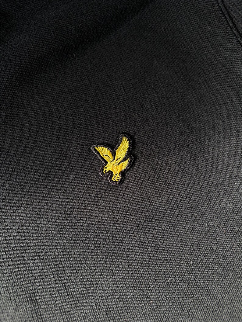 Puede incluir: Una prenda de tela negra con un peque&ntilde;o logotipo bordado de un &aacute;guila de color amarillo dorado situado en el centro. El bordado muestra un ave detallada en vuelo con las alas extendidas, realizado con hilo amarillo brillante sobre el fondo.