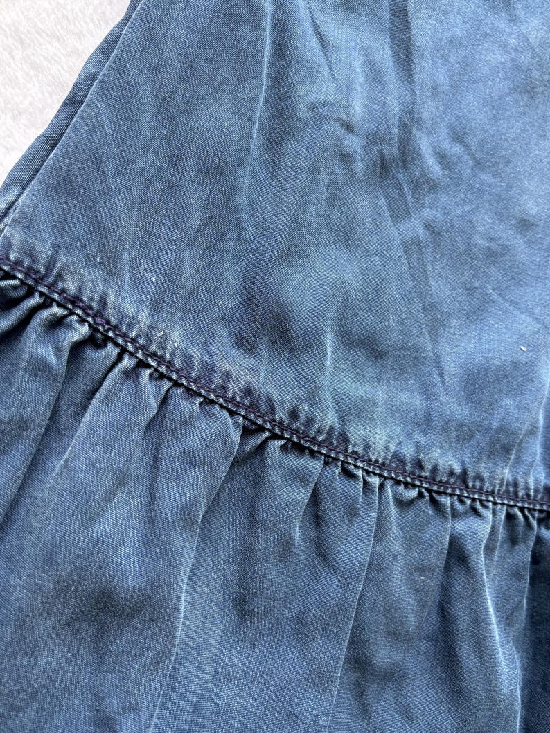 Puede incluir: Primer plano de una tela vaquera azul con un dobladillo con volantes. La tela tiene un aspecto descolorido con textura visible y sutiles variaciones de color. El dobladillo est&aacute; detallado con costuras azul oscuro.