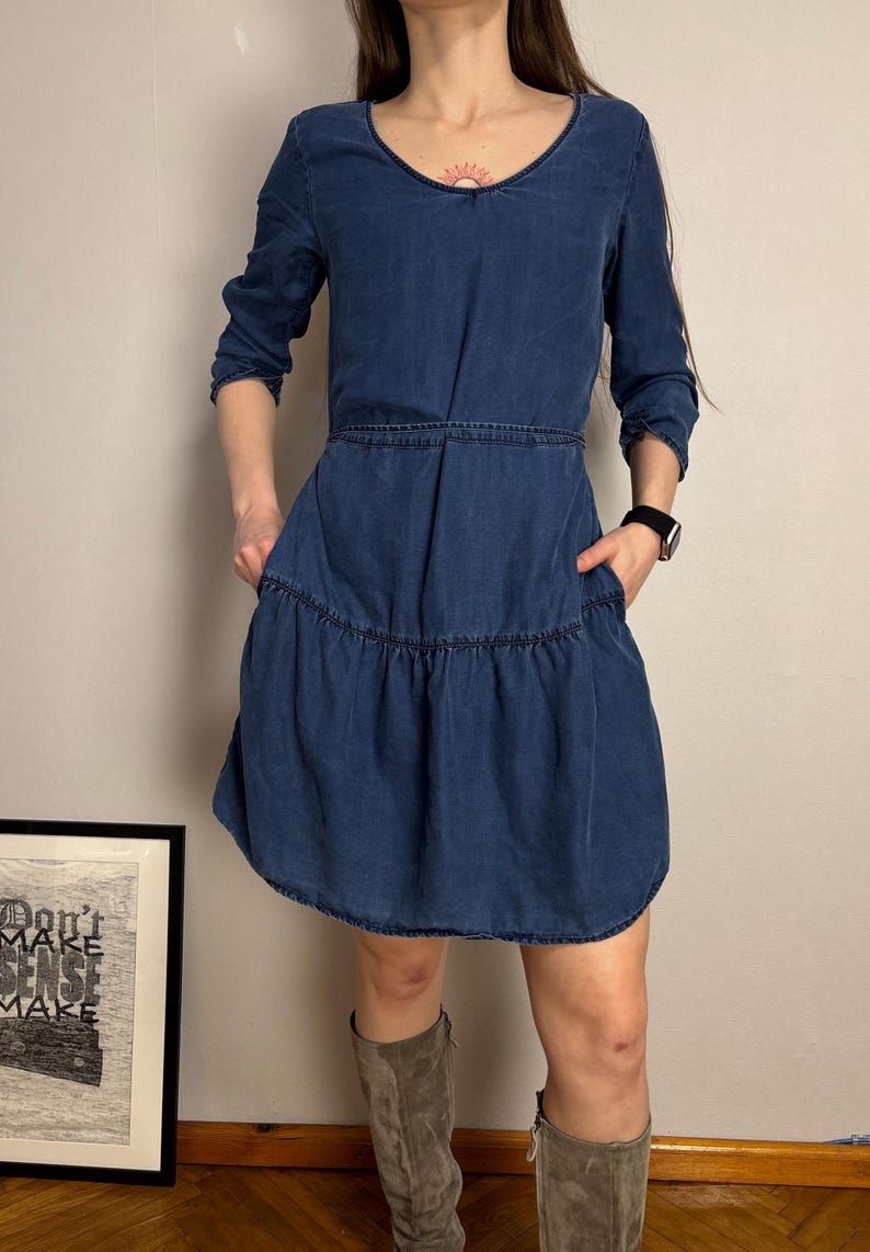 Puede incluir: Un vestido de mezclilla azul con cuello en V y mangas tres cuartos. El vestido presenta una falda escalonada y bolsillos. La persona lleva botas grises y un reloj negro. Se ve una impresi&oacute;n enmarcada con el texto "Don't Make Sense Make".