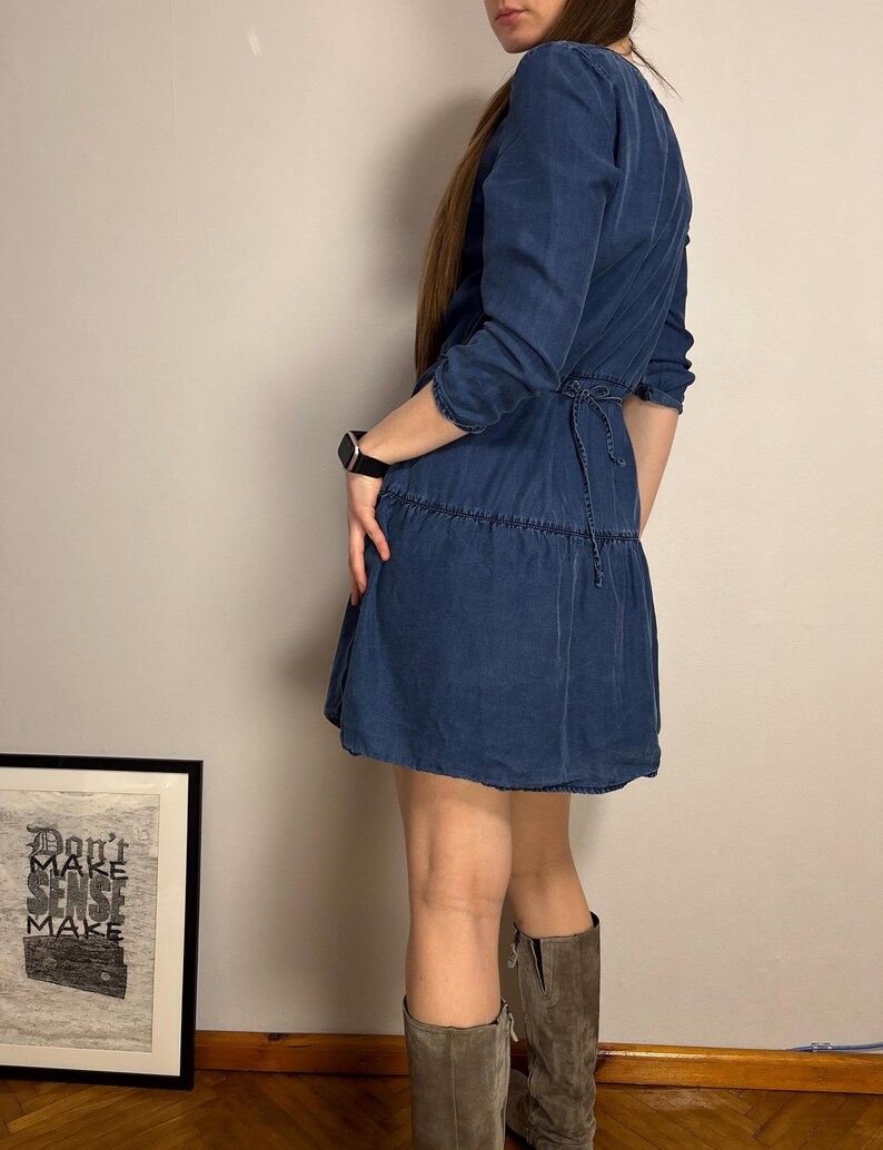 Puede incluir: Un vestido azul denim con falda escalonada y mangas tres cuartos. El vestido tiene un lazo en la cintura y se combina con botas de ante grises. En el fondo se ve una impresi&oacute;n enmarcada con el texto "Don't Make Sense Make".