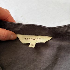 May include: A dark gray garment featuring a cream-colored interior neck label with the brand name sandwich_ printed in black text. A smaller, separate cream-colored tag attached below the main label displays the size number 36 in black.