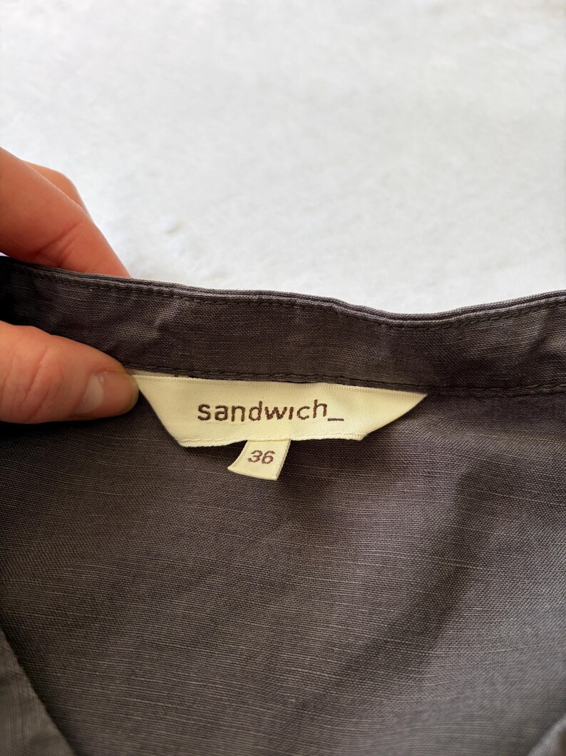 May include: A dark gray garment featuring a cream-colored interior neck label with the brand name sandwich_ printed in black text. A smaller, separate cream-colored tag attached below the main label displays the size number 36 in black.