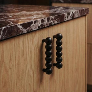 Può includere: Primo piano di un mobile da cucina con piano in marmo rosso scuro e ante in legno chiaro. Le ante dell'armadio hanno maniglie nere a perline. L'estetica generale è moderna e minimalista.