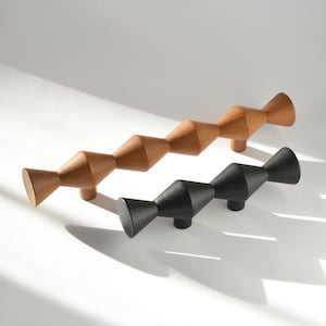 May include: Two elongated cabinet pulls, one crafted from natural light-toned wood and the other finished in a matte black. Each pull features a repeating geometric spindle shape with three distinct pointed sections and two mounting posts.