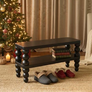 May include: A black, two-tiered bench with turned legs, holding books on the lower shelf. Two pairs of slippers, one gray and one red, sit on the woven rug in front of the bench. A decorated Christmas tree is in the background.