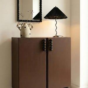 May include: A brown cabinet with two doors, black sphere feet, and black sphere door pulls. A black-framed mirror and a black lamp with a pleated shade sit on top of the cabinet. A white vase with black polka dots is also on the cabinet.