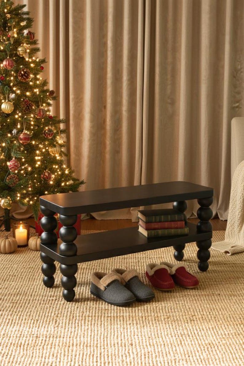 May include: A black, two-tiered bench with turned legs, holding books on the lower shelf. Two pairs of slippers, one gray and one red, sit on the woven rug in front of the bench. A decorated Christmas tree is in the background.