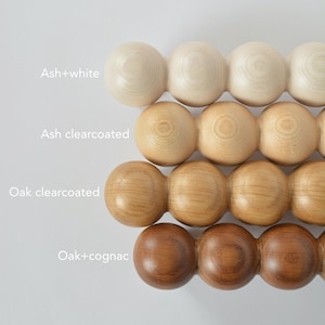 May include: Close-up of four rows of wooden massage tools in various finishes: ash+white, ash clearcoated, oak clearcoated, and oak+cognac. Each tool is composed of connected wooden spheres. Text labels identify each finish.