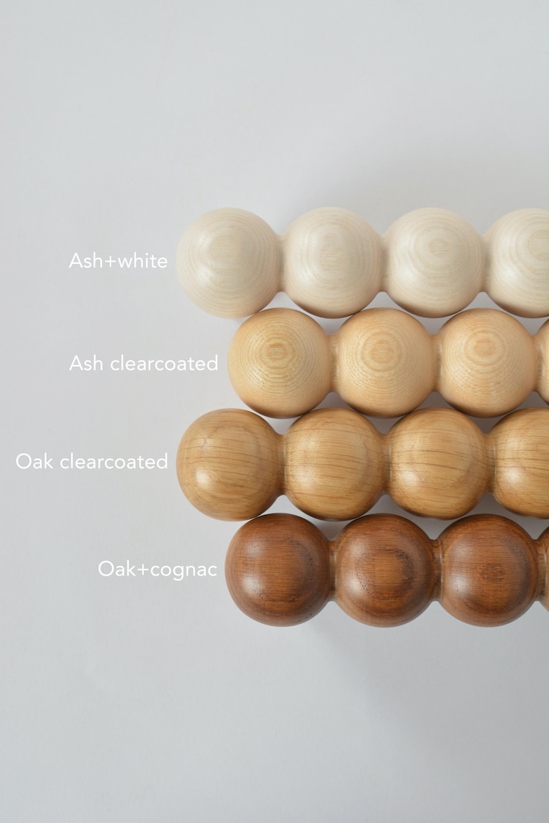 May include: Close-up of four rows of wooden massage tools in various finishes: ash+white, ash clearcoated, oak clearcoated, and oak+cognac. Each tool is composed of connected wooden spheres. Text labels identify each finish.